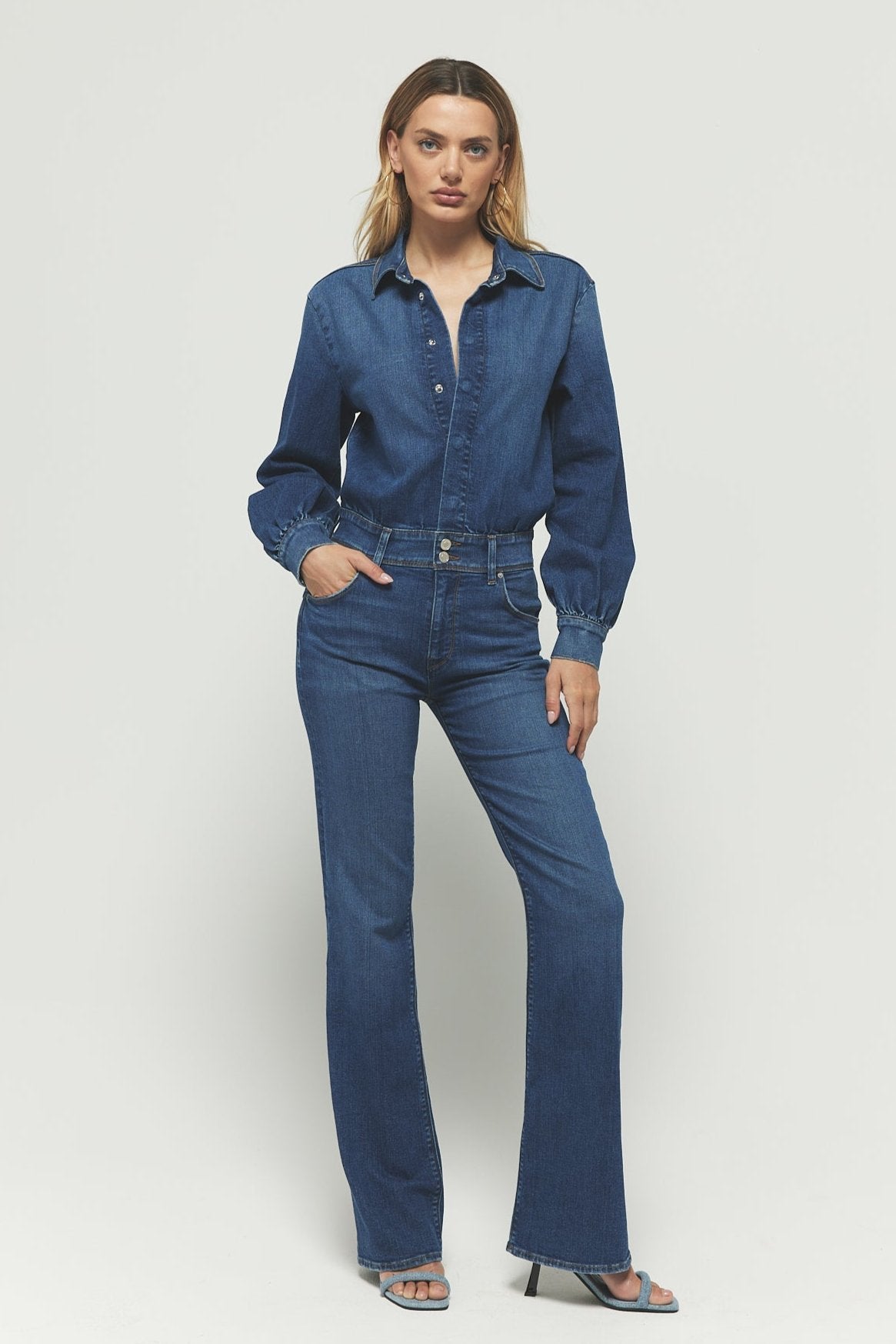 A person in a Rivet Utility The Designer denim jumpsuit poses with one hand in their pocket. The premium stretch denim features a button-up top and flared legs. Against a plain white background, the look is completed with open-toed heels.