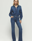 A person in a Rivet Utility The Designer denim jumpsuit poses with one hand in their pocket. The premium stretch denim features a button-up top and flared legs. Against a plain white background, the look is completed with open-toed heels.