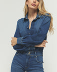 A person with long hair is confidently posing against a plain backdrop, wearing The Designer from Rivet Utility, featuring a denim shirt and high-waisted premium stretch denim jeans.